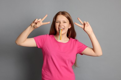 Teenage girl eating tasty rainbow sour belt while showing v-sign on grey background