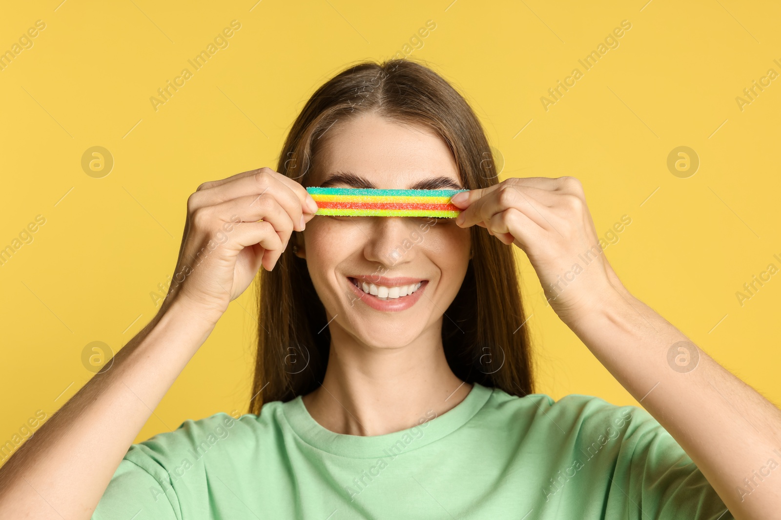 Photo of Young woman closing her eyes with tasty rainbow sour belt on yellow background