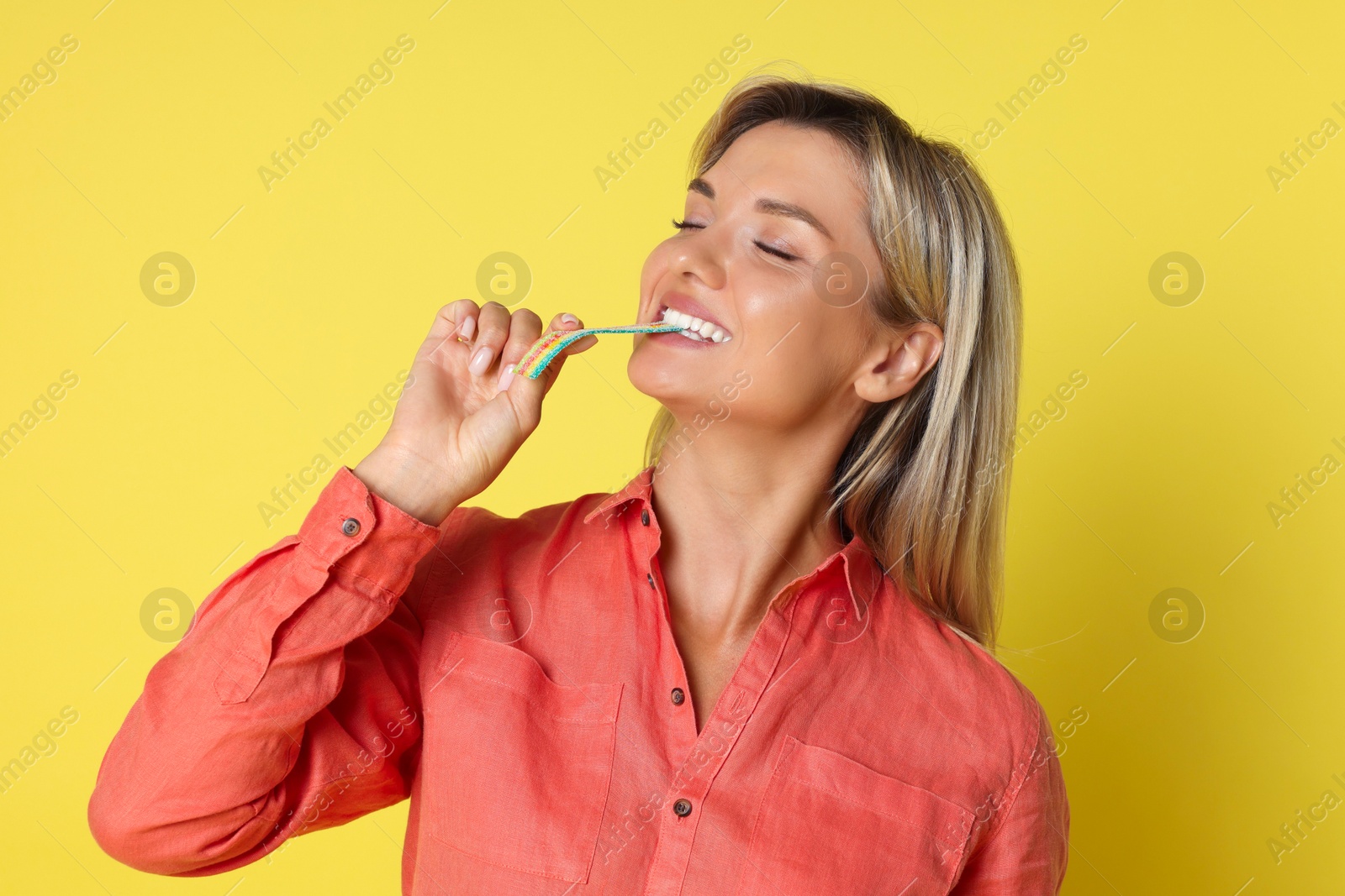 Photo of Young woman eating tasty rainbow sour belt on yellow background