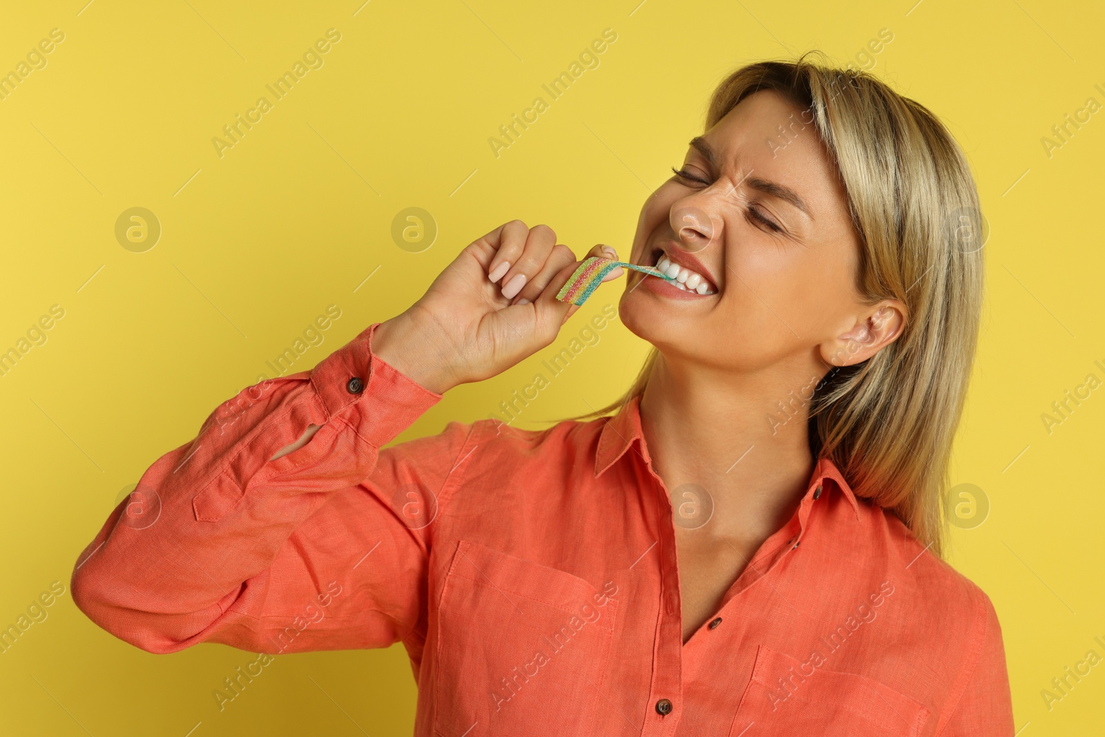 Photo of Young woman eating tasty rainbow sour belt on yellow background