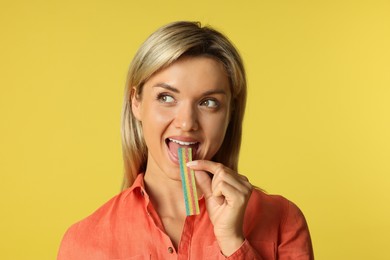 Photo of Young woman eating tasty rainbow sour belt on yellow background