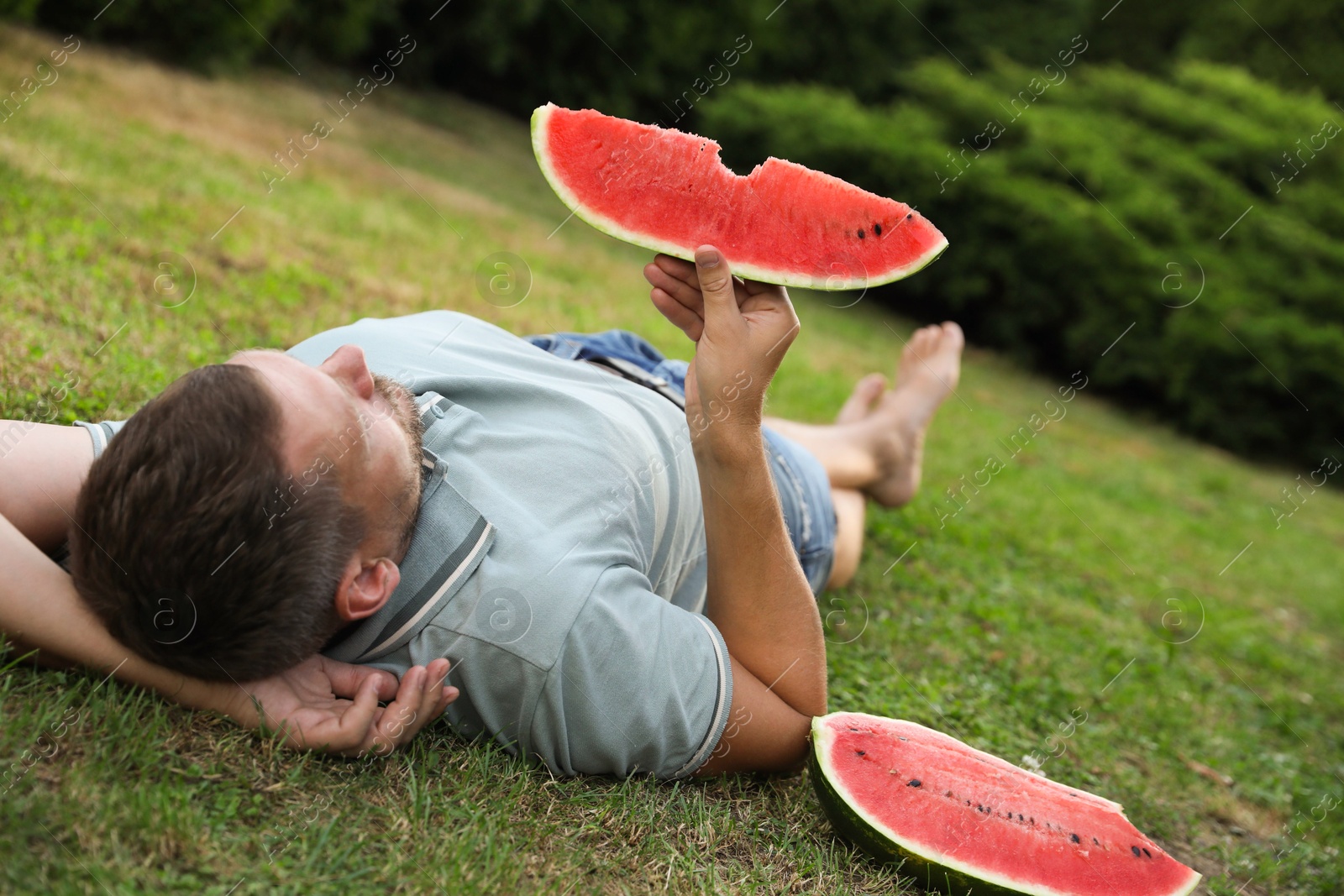 Photo of Happy man with slice of juicy watermelon on green grass outdoors