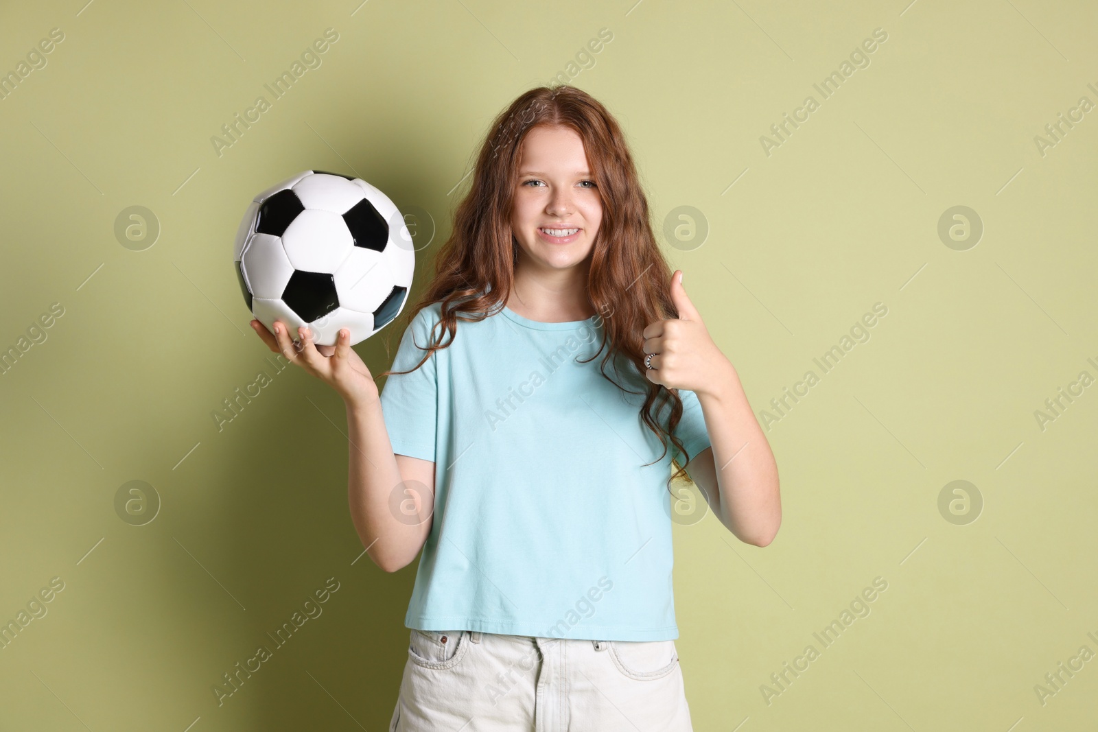 Photo of Smiling teenage girl with soccer ball showing thumbs up on green background