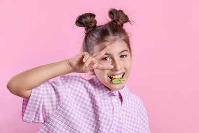 Happy girl eating tasty rainbow sour belt and showing peace sign on pink background