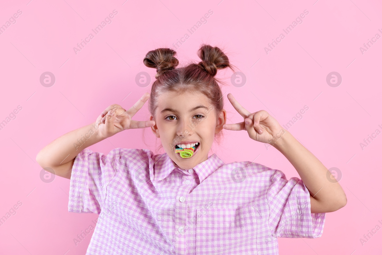 Photo of Happy girl eating tasty rainbow sour belt and showing peace signs on pink background