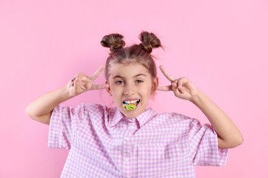 Happy girl eating tasty rainbow sour belt and showing peace signs on pink background