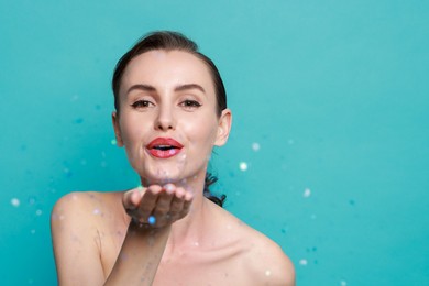 Woman with perfect makeup blowing confetti on light blue background, space for text