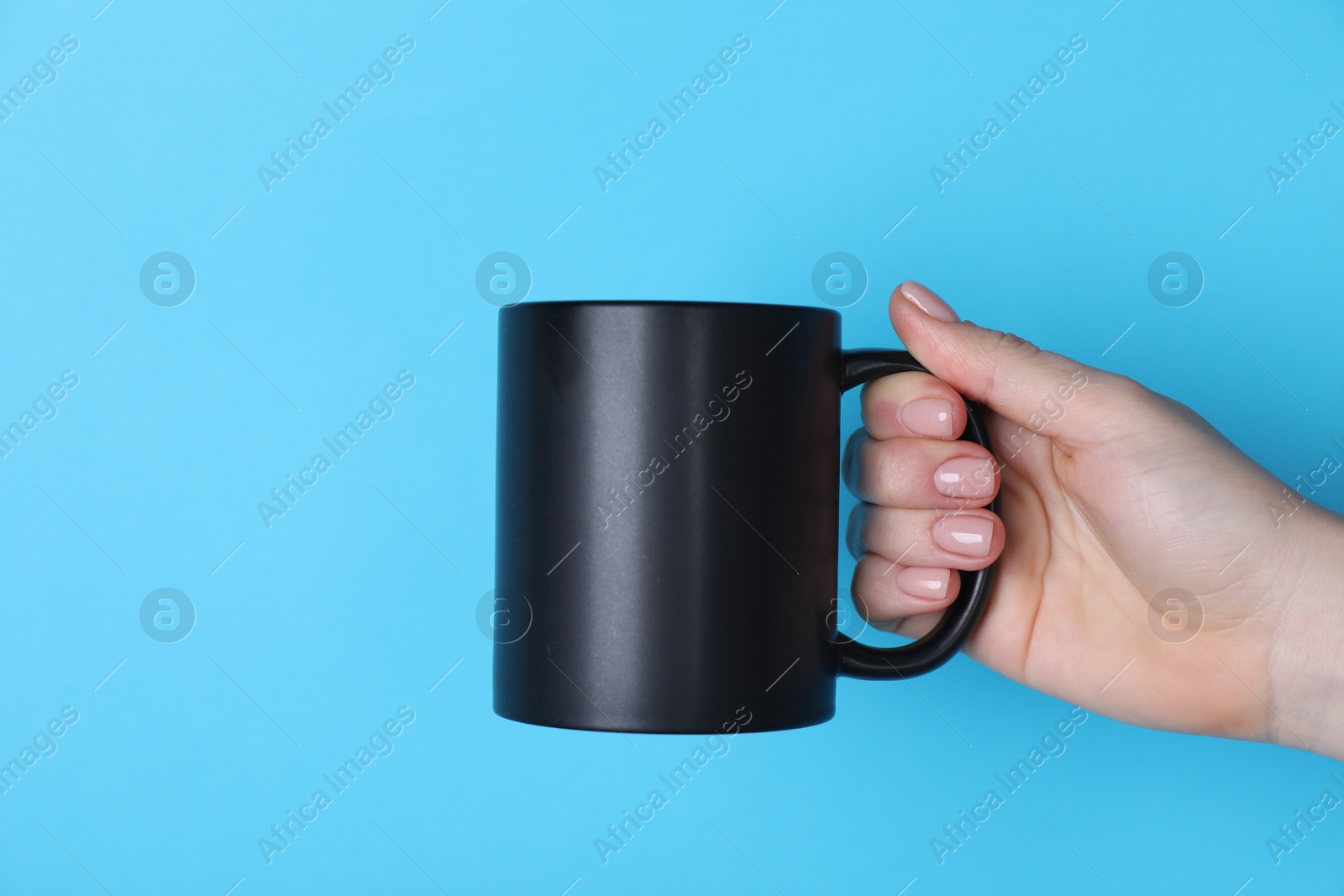 Photo of Woman holding blank black mug on light blue background, closeup. Mockup for design