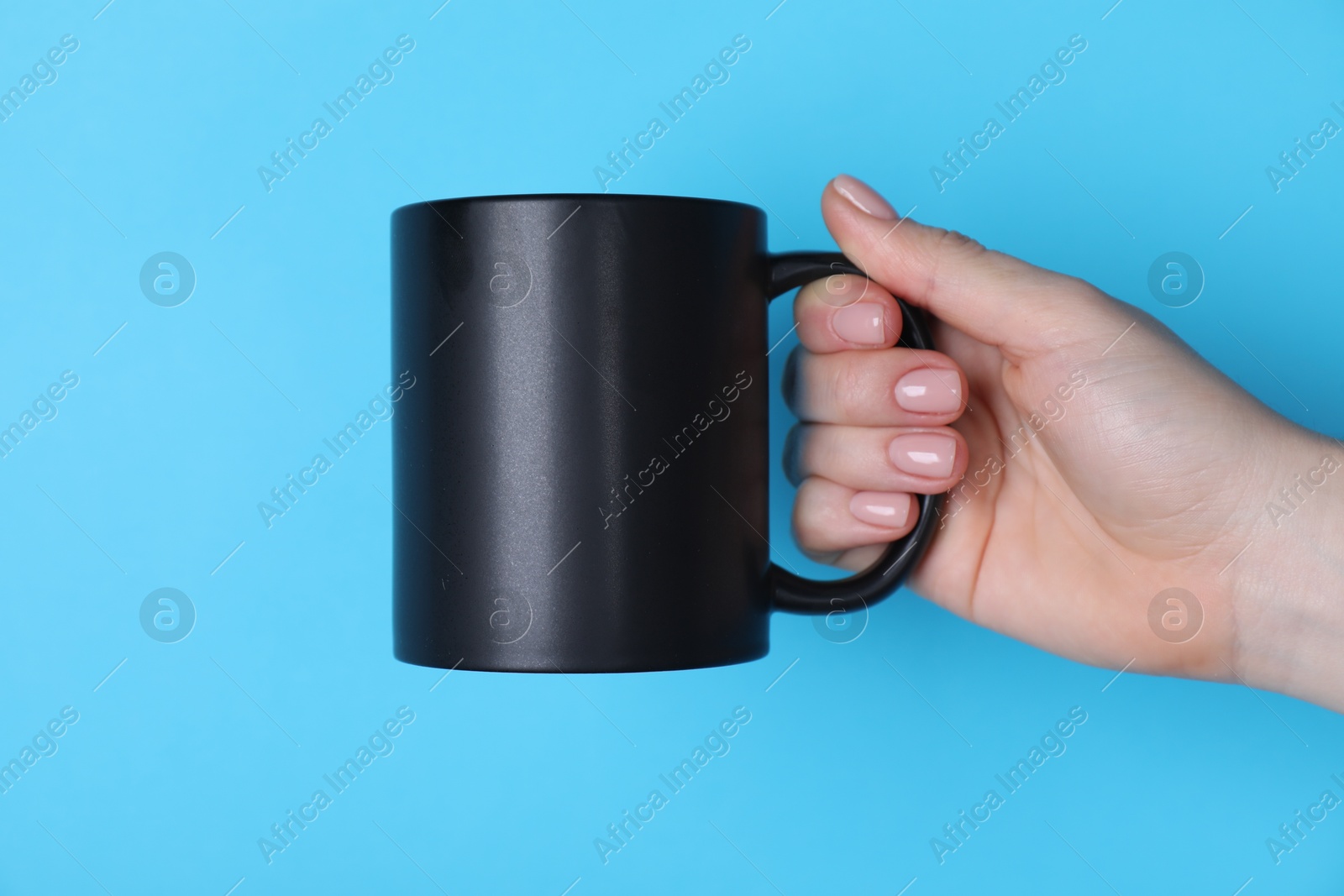 Photo of Woman holding blank black mug on light blue background, closeup. Mockup for design