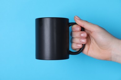 Photo of Woman holding blank black mug on light blue background, closeup. Mockup for design