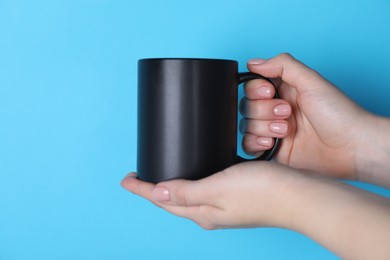 Photo of Woman holding blank black mug on light blue background, closeup. Mockup for design