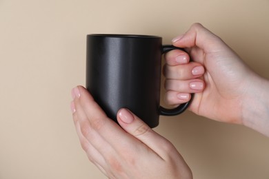 Photo of Woman holding blank black mug on beige background, closeup. Mockup for design