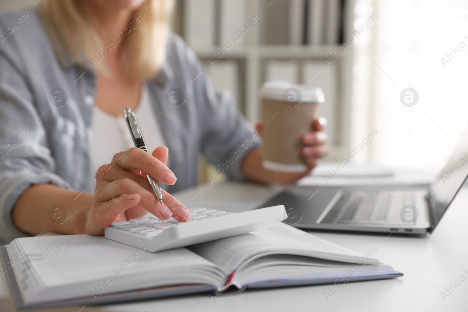 Photo of Budget planning. Woman with paper cup of coffee using calculator at table indoors, closeup