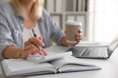 Photo of Budget planning. Woman with paper cup of coffee using calculator at table indoors, closeup