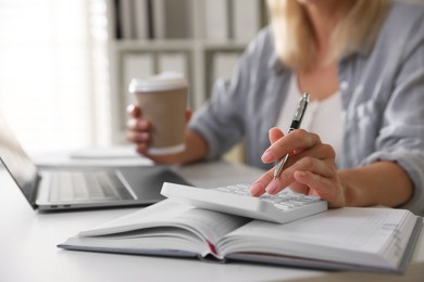 Photo of Budget planning. Woman with paper cup of coffee using calculator at table indoors, closeup