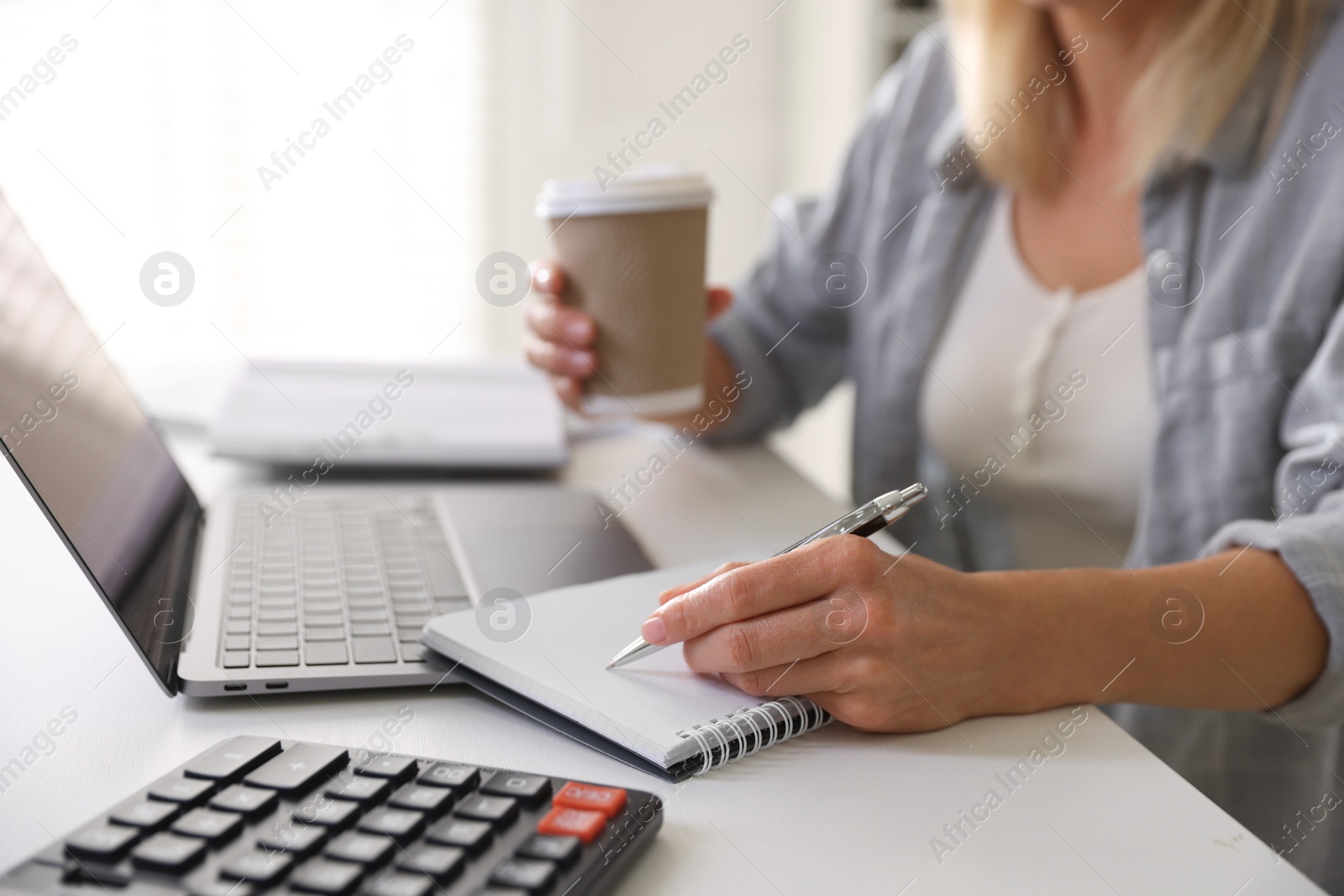 Photo of Budget planning. Woman with paper cup of coffee taking notes at table indoors, closeup