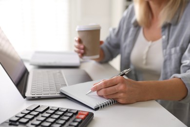 Photo of Budget planning. Woman with paper cup of coffee taking notes at table indoors, closeup