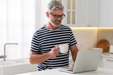 Photo of Happy middle aged man with cup of drink using laptop at white marble table in kitchen