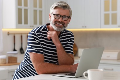 Photo of Happy middle aged man using laptop at white marble table in kitchen