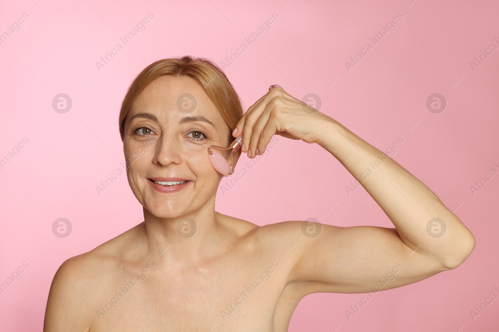 Photo of Smiling woman doing facial self massage with roller on pink background