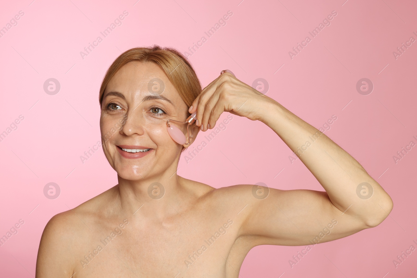 Photo of Smiling woman doing facial self massage with roller on pink background