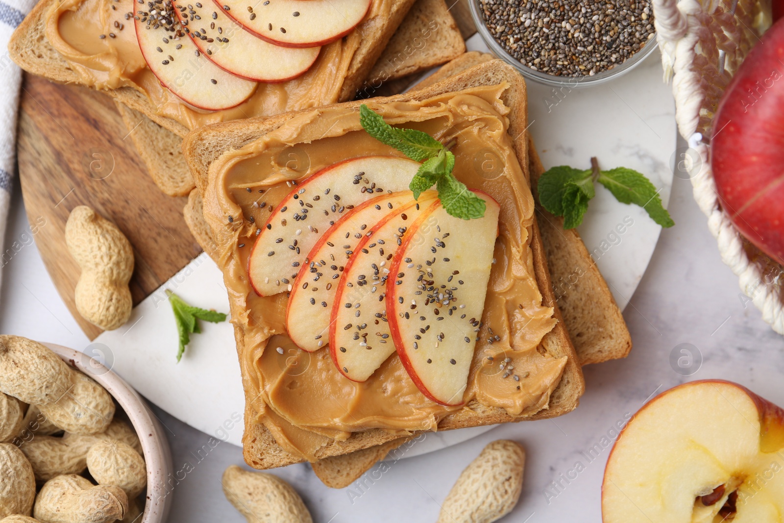 Photo of Tasty sandwiches with peanut butter, apples, chia seeds, nuts and mint on white marble table, flat lay