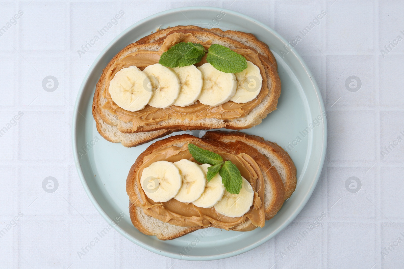 Photo of Tasty sandwiches with peanut butter, banana and mint on white tiled table, top view