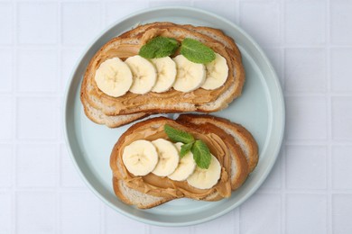 Photo of Tasty sandwiches with peanut butter, banana and mint on white tiled table, top view