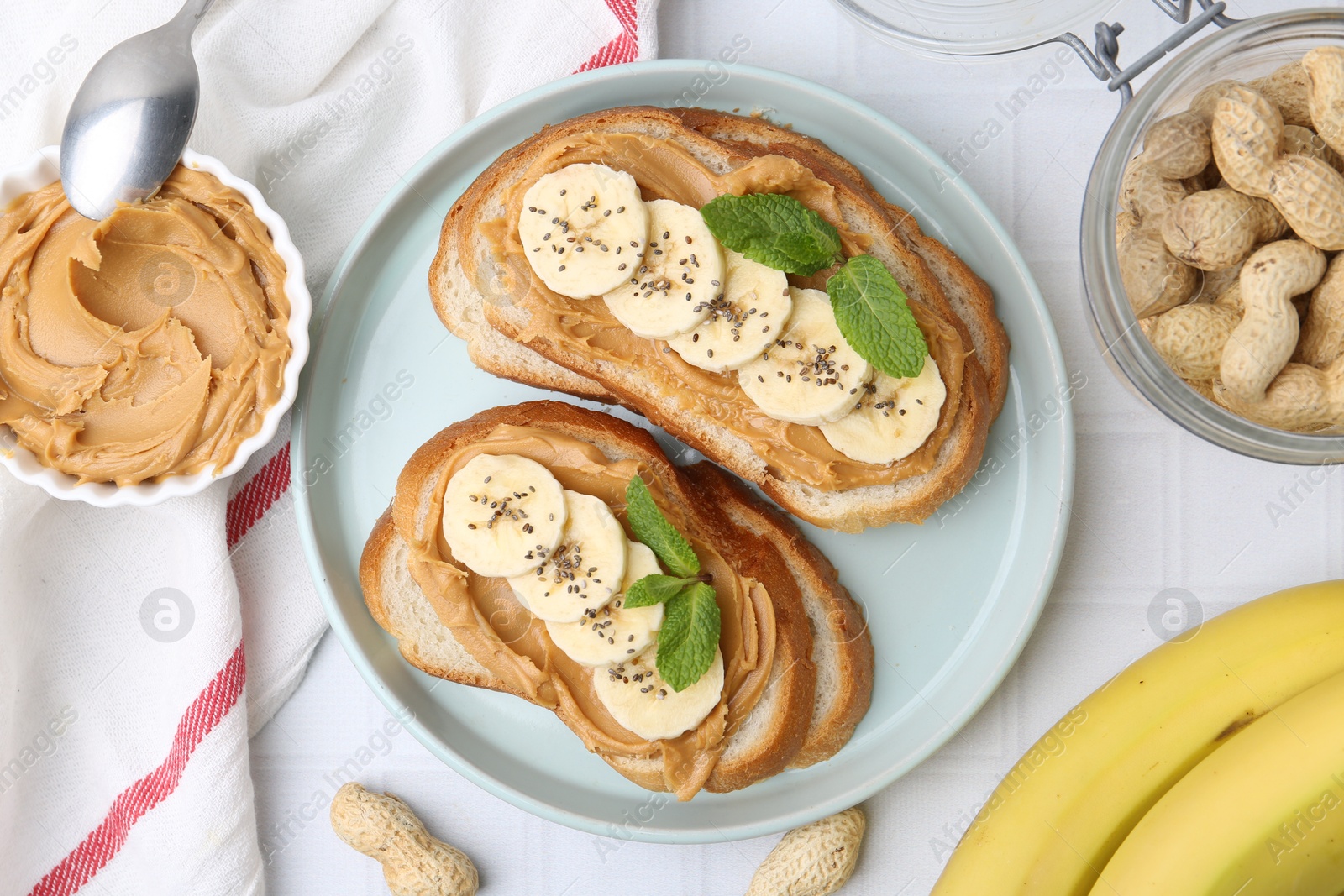 Photo of Tasty sandwiches with peanut butter, banana, chia seeds, nuts and mint on white tiled table, flat lay