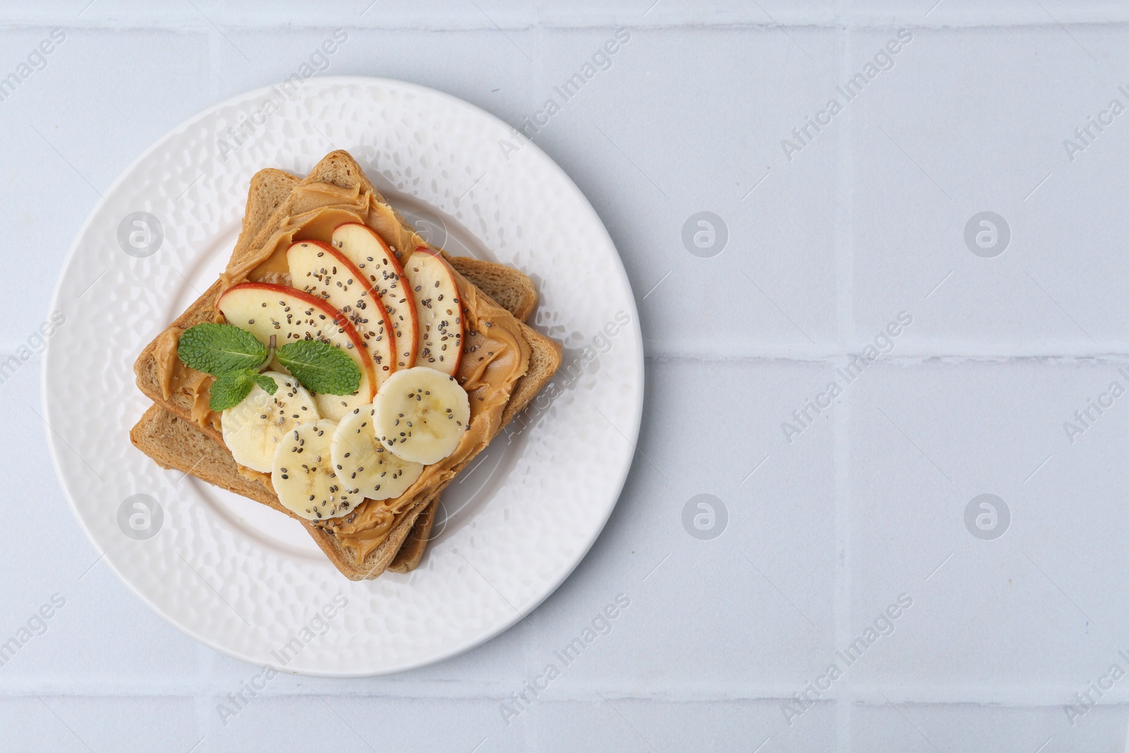 Photo of Tasty sandwich with peanut butter, apple, banana, chia seeds and mint on white tiled table, top view. Space for text