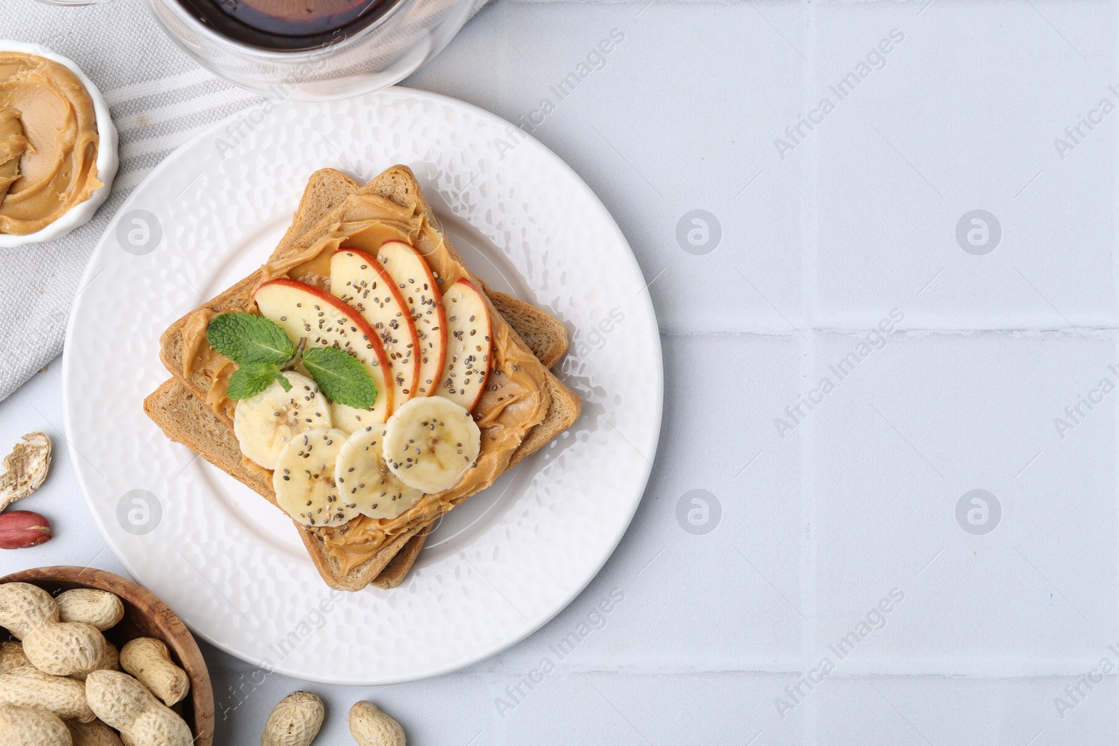 Photo of Tasty sandwich with peanut butter, apple, banana, chia seeds, nuts and mint on white tiled table, flat lay. Space for text