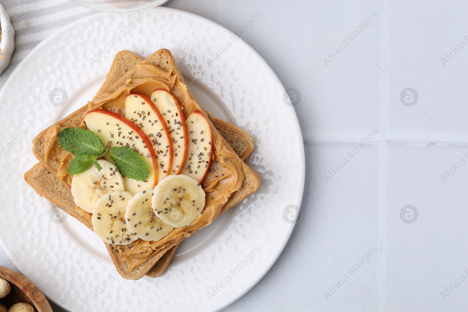 Photo of Tasty sandwich with peanut butter, apple, banana, chia seeds and mint on white tiled table, top view. Space for text