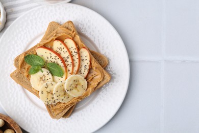 Photo of Tasty sandwich with peanut butter, apple, banana, chia seeds and mint on white tiled table, top view. Space for text