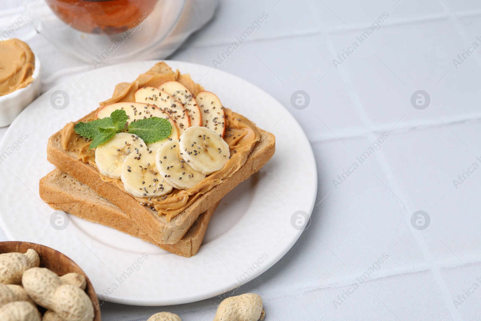 Photo of Tasty sandwich with peanut butter, apple, banana, chia seeds, nuts and mint on white tiled table, closeup. Space for text