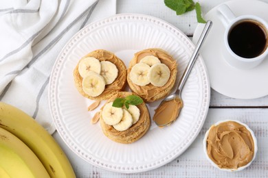 Photo of Tasty sandwiches with peanut butter, banana, mint, coffee and spoon on white wooden table, flat lay