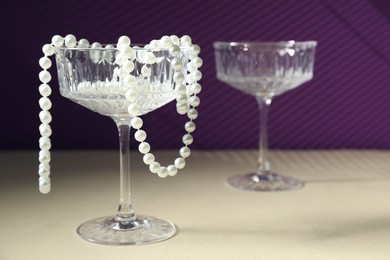 Photo of Elegant wine glasses and pearl jewelry on beige table against purple background