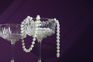 Photo of Elegant wine glasses and pearl jewelry on purple background, space for text