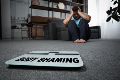 Stop body shaming. Depressed overweight man and floor scales indoors, selective focus