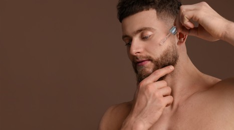 Young man applying serum on face against brown background. Banner design with space for text. Skin care