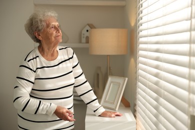 Photo of Loneliness concept. Sad senior woman looking through window blinds at home