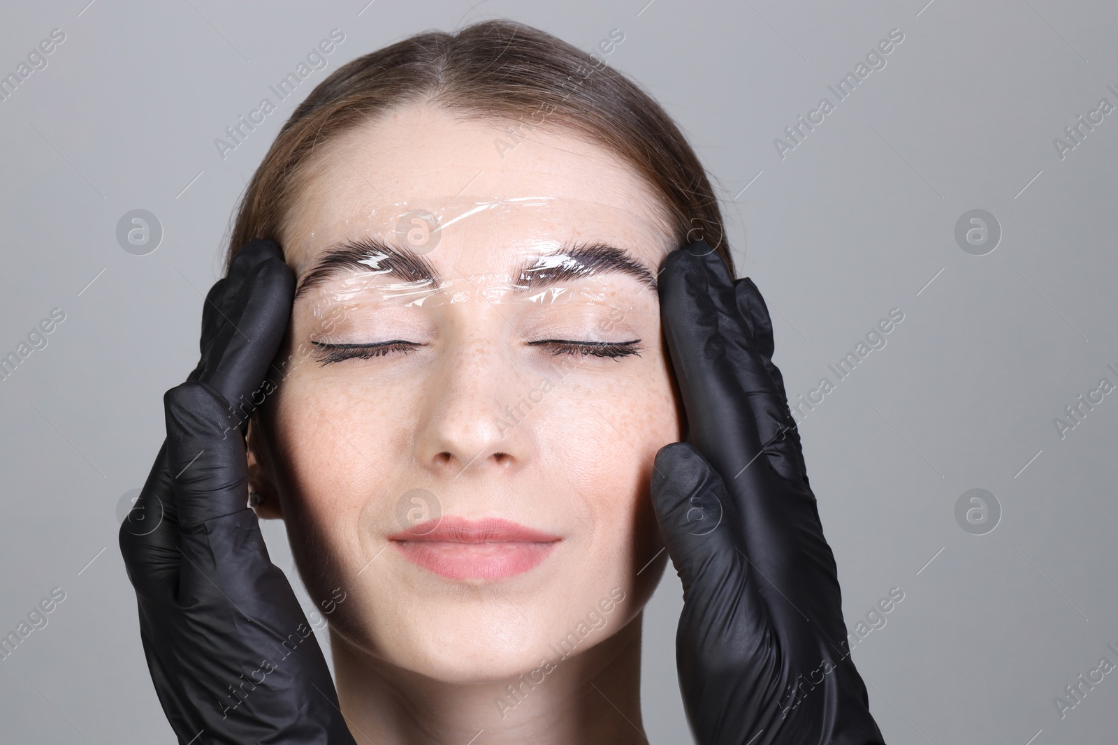 Photo of Brow lamination. Cosmetologist putting film onto woman's eyebrows against grey background, closeup