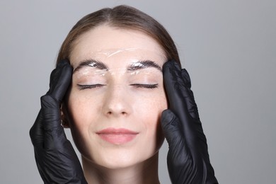 Brow lamination. Cosmetologist putting film onto woman's eyebrows against grey background, closeup