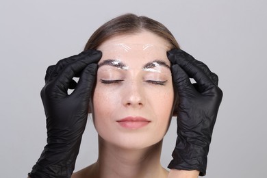 Photo of Brow lamination. Cosmetologist putting film onto woman's eyebrows against grey background, closeup