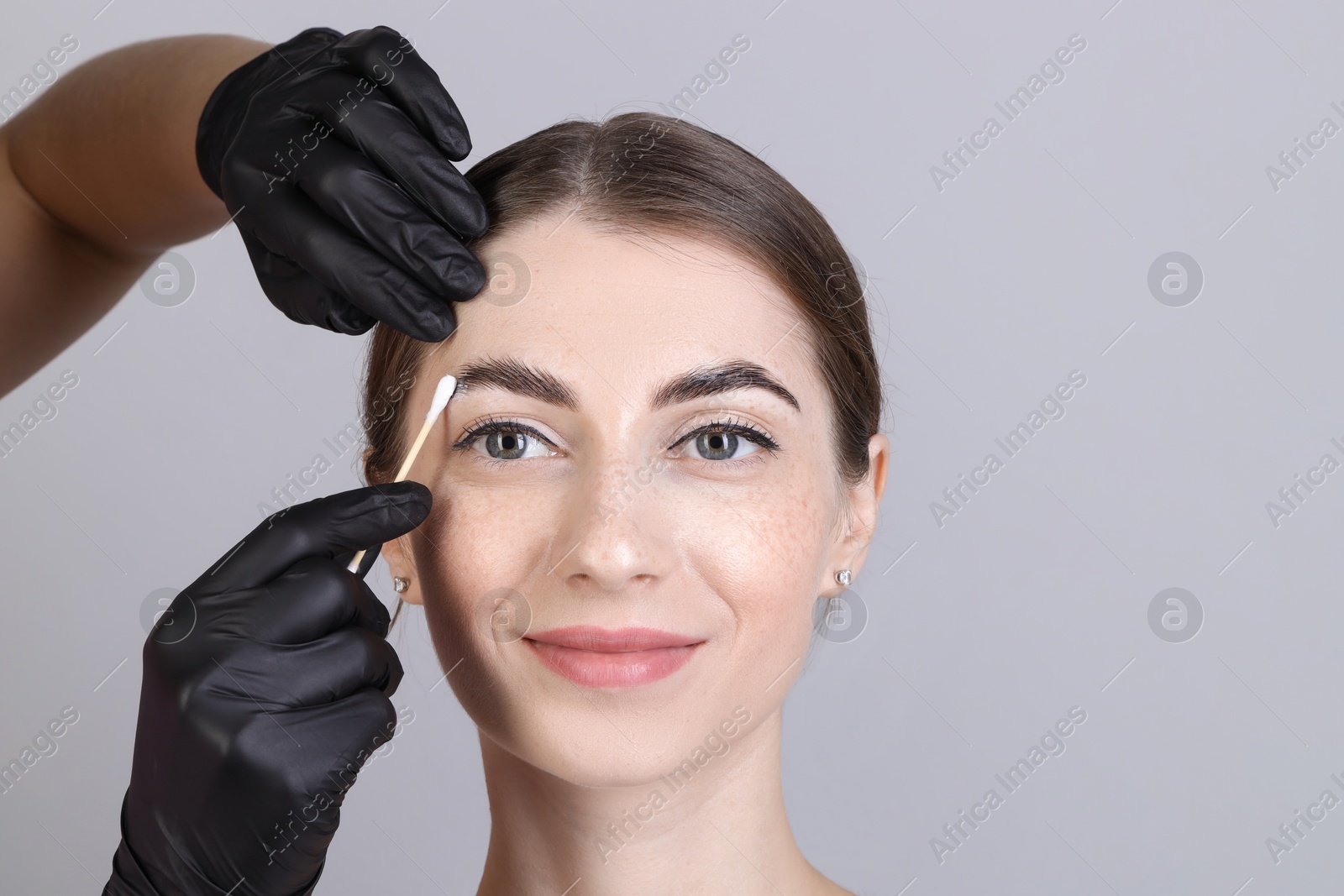 Photo of Brow lamination. Cosmetologist applying cream onto woman's eyebrows against grey background, closeup