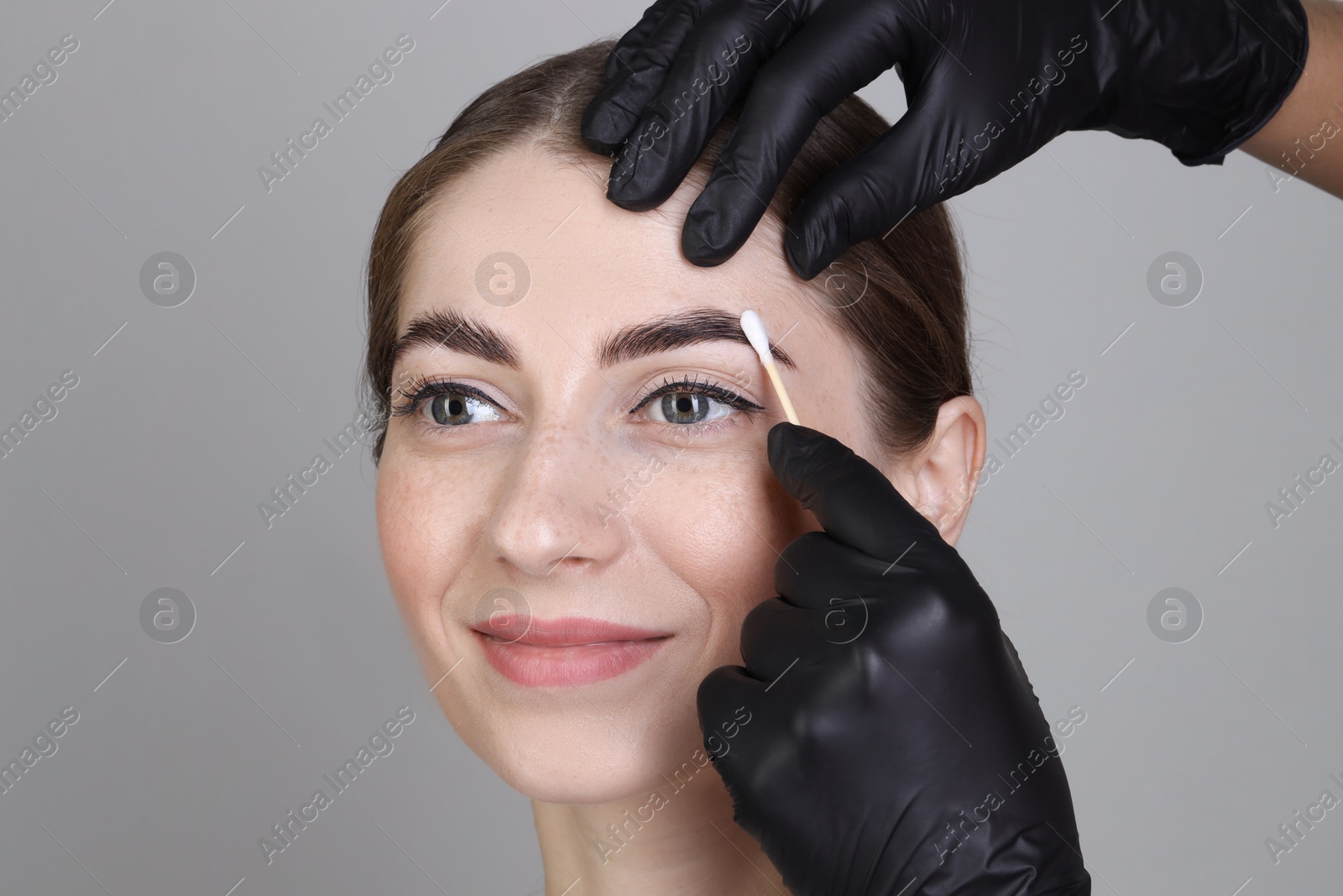 Photo of Brow lamination. Cosmetologist applying cream onto woman's eyebrows against grey background, closeup