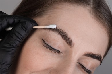 Photo of Brow lamination. Cosmetologist applying cream onto woman's eyebrows against grey background, closeup