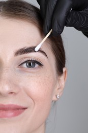 Photo of Brow lamination. Cosmetologist applying cream onto woman's eyebrows against grey background, closeup