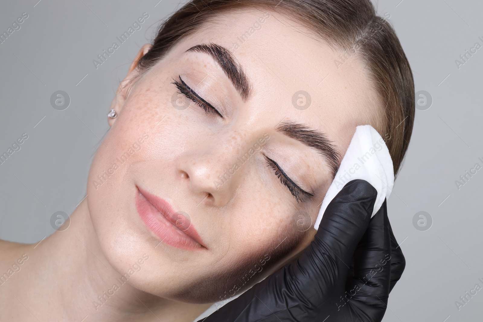 Photo of Brow lamination. Cosmetologist wiping woman's eyebrows with cotton pad against grey background, closeup