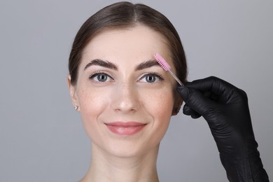 Photo of Brow lamination. Cosmetologist combing woman's eyebrows with brush against grey background, closeup