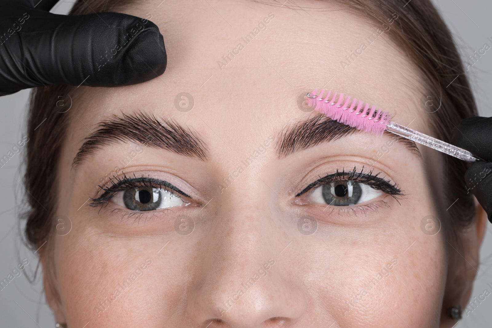 Photo of Brow lamination. Cosmetologist combing woman's eyebrows with brush against grey background, closeup